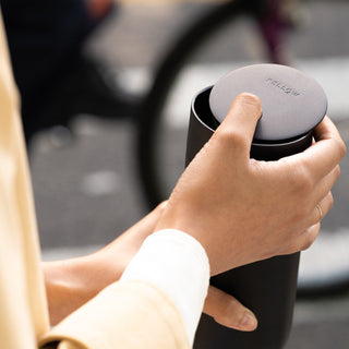 Hand holding black FELLOW Carter Slide Mug 16oz with textured lid outdoors on the street