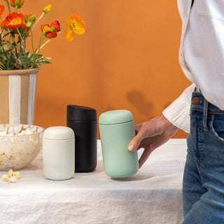 FELLOW Carter Slide Mug 12oz in three colors on white table with orange background and hand reaching