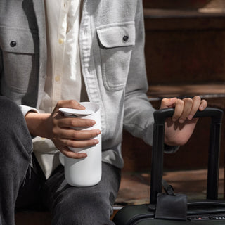 FELLOW Carter Slide Mug 12oz in white held by person sitting on steps next to black suitcase handle