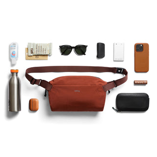 Bellroy Lite Sling in rust color with water-resistant fabric and adjustable strap surrounded by essentials
