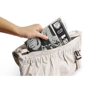 Hand placing newspaper into beige Bellroy Cinch Messenger Bag pocket with metal buckle closure