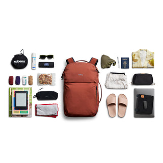 Bellroy Lite Travel Pack 30L in rust red with travel essentials flat lay including apparel and accessories