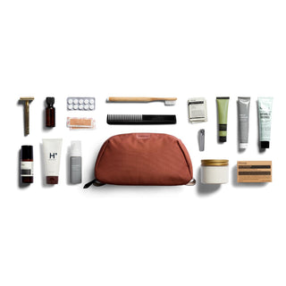 Bellroy Toiletry Kit Plus rust fabric pouch with assorted personal care items neatly arranged on white background