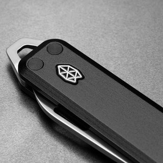 James The Elko Knife showing black Micarta handle with metal emblem and stainless steel clip on gray surface
