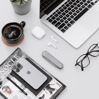 James The Wells Knife in gray aerospace aluminum placed among tech and office accessories on a white desk surface