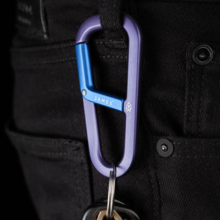 James The Hardin Carabiner in anodized purple and blue aluminum clipped onto black fabric loop