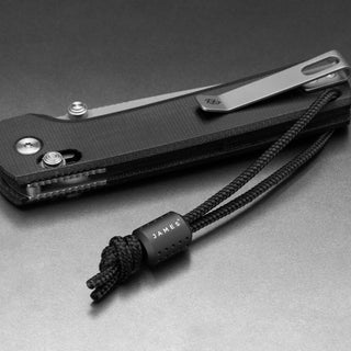 James The Alder Lanyard & Bead in black nylon paracord with anodized black bead next to a folding knife