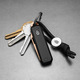 James The Elko Knife with black G10 handle attached to keyring and black paracord lanyard on gray surface
