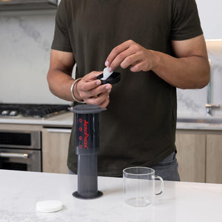 Person placing AeroPress Microfilters (350-Pack) into black coffee maker filter cap on kitchen counter