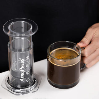 AeroPress Flow Control Filter Cap installed on clear plastic coffee brewer beside glass cup of black coffee