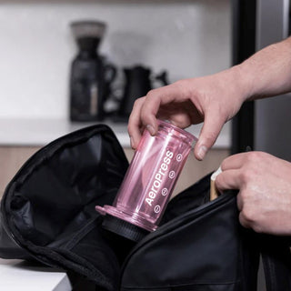 A person placing a pink AeroPress Clear Coffee Maker into a black travel bag on a kitchen counter