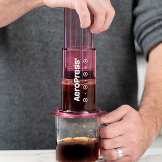 AeroPress Clear Coffee Maker in translucent purple pressing freshly brewed coffee into a glass mug