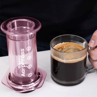 AeroPress Clear Coffee Maker with translucent purple brewing chamber next to a clear glass mug of black coffee
