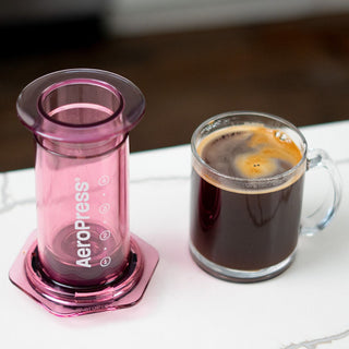 AeroPress Clear Coffee Maker in translucent pink next to a glass mug with black coffee and crema