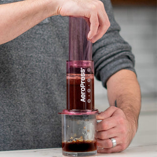 AeroPress Clear Coffee Maker in purple brewing fresh coffee over a glass mug on countertop