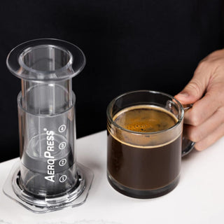 AeroPress Clear Coffee Maker next to a hand holding a glass mug filled with freshly brewed coffee
