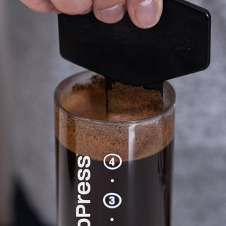 AeroPress Clear Coffee Maker showing coffee grounds being pressed with black plunger tool close-up