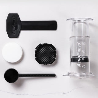 AeroPress Clear Coffee Maker with plunger, filter cap, scoop, and stirrer arranged on white surface