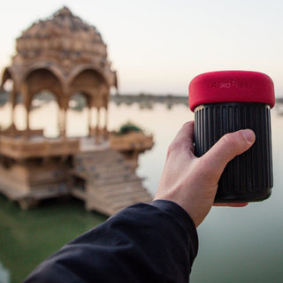 Hand holding AeroPress Go Coffee Maker with textured black body and red lid near lakeside pavilion at sunset