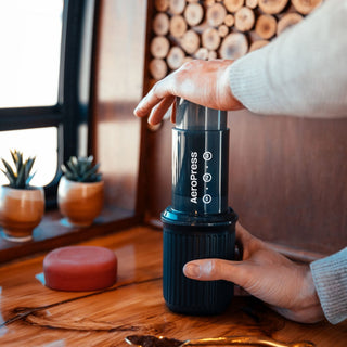 Hands pressing down the black AeroPress Go Coffee Maker on a wooden surface near potted plants and window light