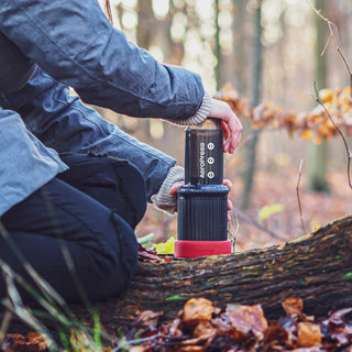 A person using AeroPress Go Coffee Maker outdoors on a tree trunk in a forest setting