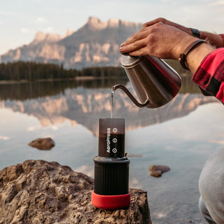 AeroPress Go Coffee Maker with black and red body brewing coffee outdoors by a lake at sunrise