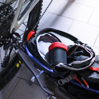 AeroPress Go Coffee Maker with red lid packed inside a black and blue cycling backpack near bicycle wheel