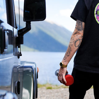 Person holding red AeroPress Go Coffee Maker near a vehicle with mountain and lake background