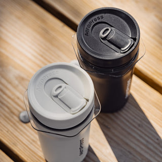 Two AeroPress Go Plus Travel System coffee makers in black and white with secure flip lids on wooden surface