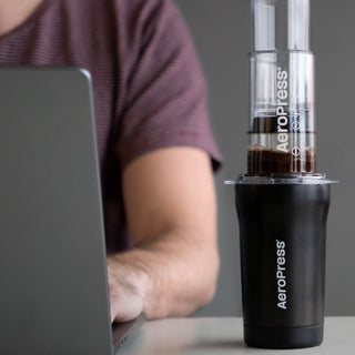 AeroPress Go Plus Travel System brewing coffee with black cup beside person using laptop