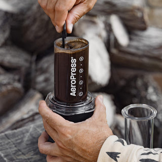 A person stirring brewed coffee in a clear AeroPress Go Plus Travel System on rustic wood background