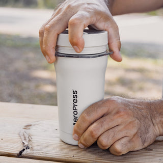 A hand holding the white AeroPress Go Plus Travel System coffee maker on a wooden table outdoors