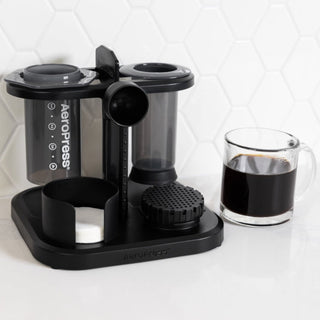AeroPress Organizer in black plastic holding coffee filters and accessories next to a glass mug of black coffee
