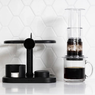 AeroPress Organizer in black holding AeroPress components beside brewing coffee cup on white surface
