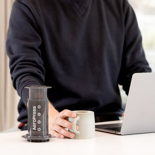 A person holding a mug beside the black AeroPress Original Coffee Maker on a white table near a laptop