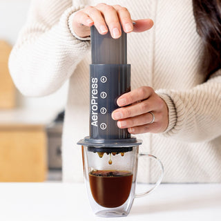 AeroPress Original Coffee Maker brewing dark coffee with clear glass cup and hand pressing top plunger