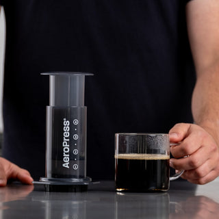 AeroPress Original Coffee Maker with clear chamber next to freshly brewed black coffee mug