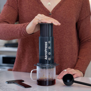 A person pressing coffee with black AeroPress Original Coffee Maker on clear glass mug