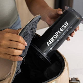 A person placing a black AeroPress XL Coffee Press with logo into a beige bag pocket close-up