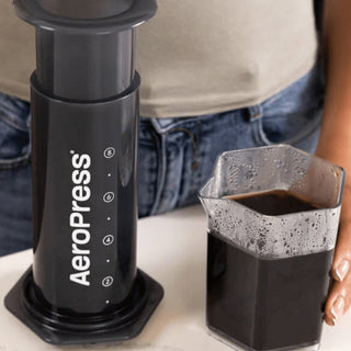 AeroPress XL Coffee Press in black with measuring markings beside a glass container of brewed coffee