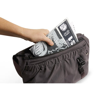 Hand placing folded newspaper into front pocket of Bellroy Cinch Messenger Bag in dark fabric
