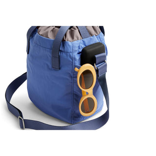 Bellroy Cinch Bucket Bag in blue recycled nylon with adjustable strap and side pocket holding sunglasses and case