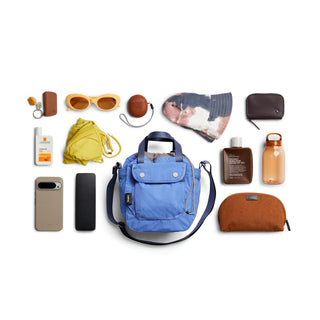Bellroy Cinch Bucket Bag in blue with front flap and adjustable shoulder strap surrounded by personal items
