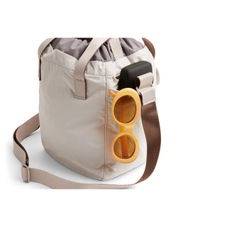 Bellroy Cinch Bucket Bag in light beige with front pocket holding sunglasses and phone strap detail