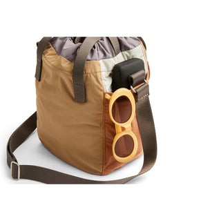 Bellroy Cinch Bucket Bag in tan with adjustable strap and side pocket holding sunglasses