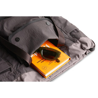 Bellroy Cinch Tote in dark gray with visible snap buttons and brown strap over an orange book and sunglasses