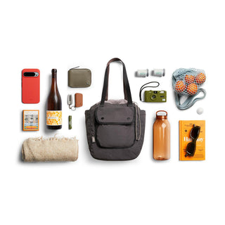 Bellroy Cinch Tote in charcoal with front pocket and shoulder straps, surrounded by everyday essentials