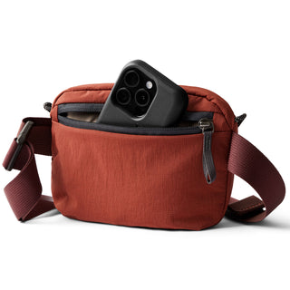 Bellroy Lite Belt Bag in rust red with front zip pocket holding a black smartphone