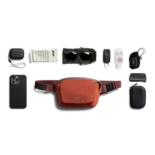 Bellroy Lite Belt Bag in rust color with compact design surrounded by everyday essentials.