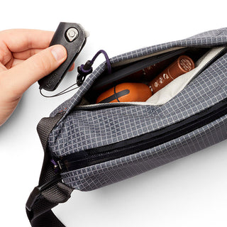 Bellroy Lite Sling Mini in textured gray fabric with hand holding a key fob near open zipper compartment
